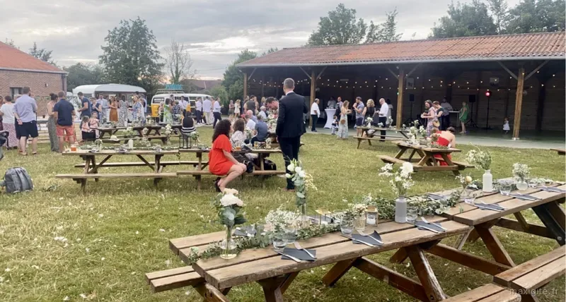 Réception en plein air sous la grange ouverte, tables en bois fleuries et invités attablés