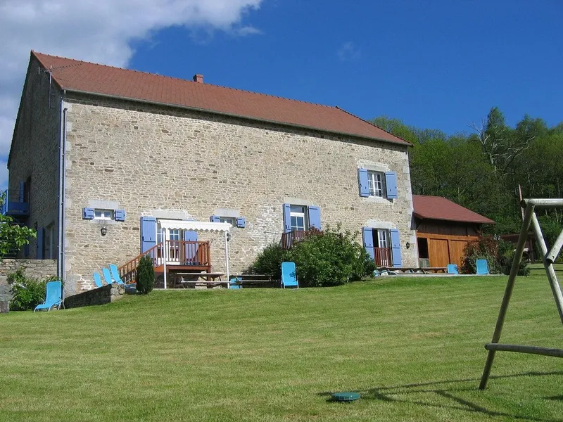 Façade en pierre du corps de ferme avec toit de tuiles rouges, volets bleus et grand jardin arboré en pente douce