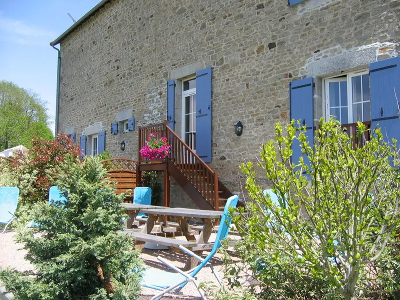 Terrasse en bois du gîte avec table de pique-nique, chaises longues bleues et façade en pierre du bâtiment
