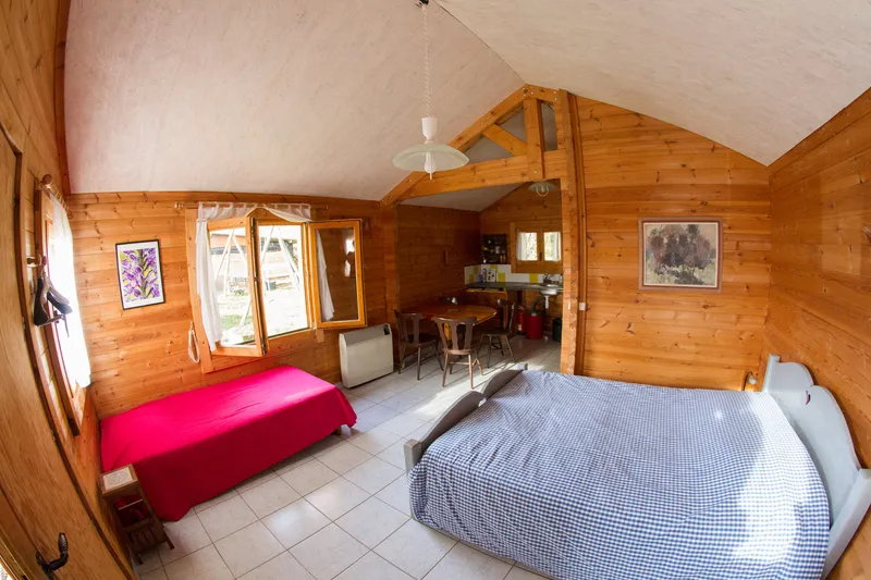 Chambre de chalet avec grand lit à carreaux bleus et lit d'appoint rouge, coin repas en bois à l'arrière-plan