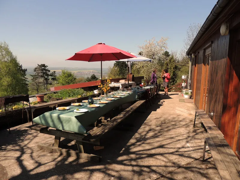 Longue tablée conviviale sous parasol rouge sur la terrasse surplombant la vallée, avec vue dégagée sur la campagne