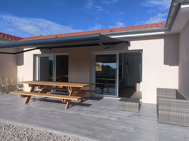 Terrasse du gite l'Ambremy avec table de pique-nique en bois, store banne deploye et baies vitrees ouvrant sur la piece de vie