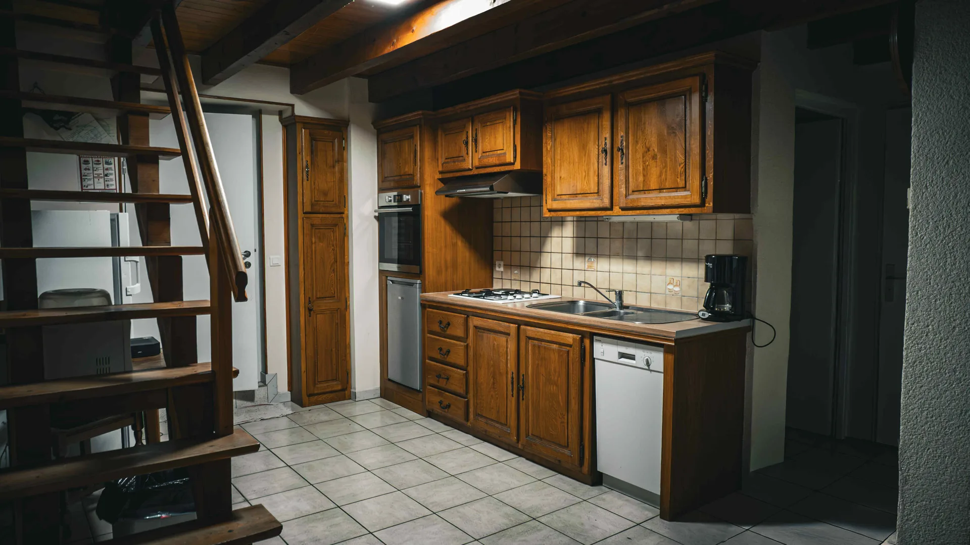 Cuisine équipée en bois avec plaque de cuisson, four encastré et lave-vaisselle, escalier visible à gauche