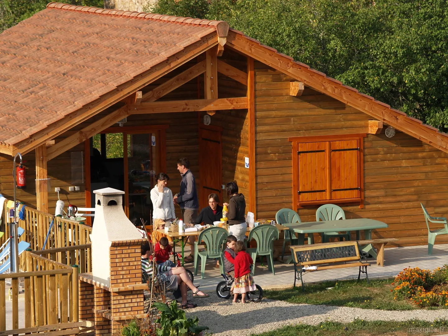 Terrasse d'un chalet en bois avec cheminée barbecue en brique et famille attablée autour d'un repas