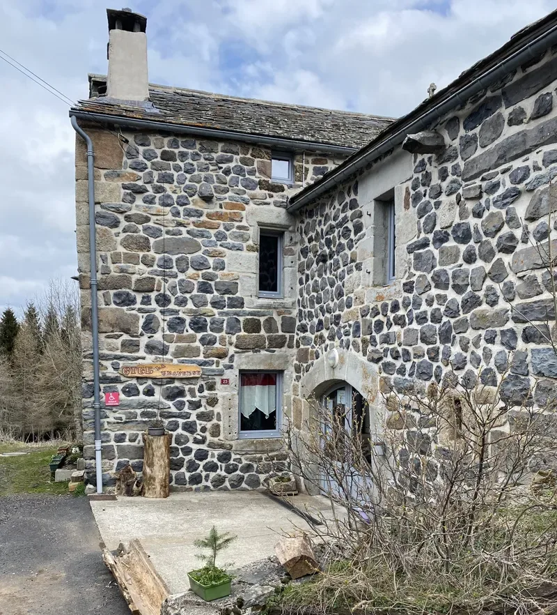 Façade en pierre du gîte avec panneau 'Gîte la Traverse', bûches empilées en décor et porte voûtée bleu ciel